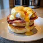 Close-up of a perfectly made eggs benedict stack with hollandaise sauce dripping over poached egg and ham on an English muffin.