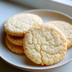 Close-up of several perfectly round, slightly crinkled sugar cookie rounds stacked on a light ceramic plate.
