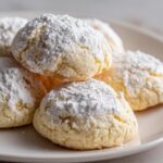 A close-up of several soft, pale yellow lemon cookies generously dusted with powdered sugar.