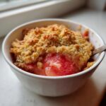 Close-up of a single serving of warm rhubarb crisp with a golden, crumbly topping in a white bowl.