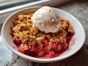 Close-up of a warm serving of rhubarb crisp with a golden oat topping and a scoop of vanilla ice cream.