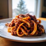 A close-up of thick pasta noodles generously coated in rich bolognese sauce on a white plate.