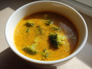 Close-up of a creamy, vibrant orange broccoli cheddar soup with visible florets in a white bowl.