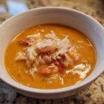 A close-up of a bowl of rich, orange seafood soup generously topped with shredded white crab meat and pieces of shrimp.
