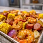 Close-up of a sheet pan filled with seasoned shrimp boil ingredients including large shrimp, corn on the cob, sausage, and onions.