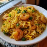 Close-up of a white bowl filled with vibrant yellow shrimp fried rice recipe, topped with pink shrimp and green onions.