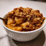 Close-up of a single serving of warm apple crisp in a white ramekin, showing caramelized apples and a crunchy oat topping.