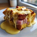 A decadent slice of eggs benedict casserole featuring layers of bread, ham, and topped generously with creamy hollandaise sauce.