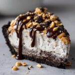 A rich slice of peanut butter ice cream pie with a chocolate crust, topped with fudge, peanuts, and cookie crumbs.