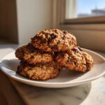 A stack of four soft banana oatmeal cookies with visible chocolate chips resting on a white plate near a window.