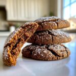 A stack of three soft gingerbread cookies, topped with sugar crystals, with one cookie broken open showing its moist interior.