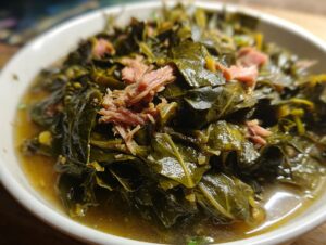 Close-up of tender, simmered Southern collard greens served in a white bowl with savory broth and pieces of shredded smoked ham.
