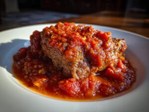 A close-up of tender steak pizzaiola smothered in a chunky, vibrant red tomato sauce on a white plate.
