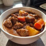 Close-up of a white bowl filled with hot, steaming crockpot beef stew featuring tender beef chunks, carrots, and potatoes.