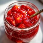 Close-up of chunky strawberry freezer jam in a glass jar with a small golden spoon resting inside.