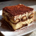 A perfect square slice of homemade tiramisu recipe layered with cream and coffee-soaked ladyfingers, dusted heavily with cocoa powder.
