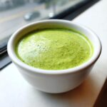 A small white bowl filled with vibrant, creamy cilantro lime sauce, sitting near a window.
