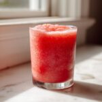Close-up of a vibrant, icy watermelon smoothie served in a clear glass on a sunny countertop.