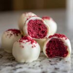 Close-up of white chocolate coated red velvet cake balls, one is broken open showing the bright red interior.
