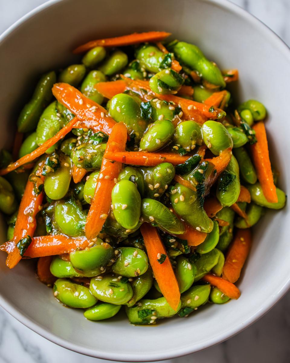 Close-up of a bright green edamame salad mixed with julienned carrots and sesame seeds in a white bowl.