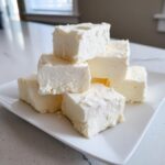 A stack of creamy, white squares of homemade white chocolate fudge resting on a white plate.