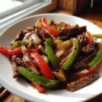 A close-up of a white bowl filled with tender pepper steak strips mixed with vibrant red and green bell peppers and onions.