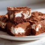 Close-up of rich chocolate fudge squares with visible white marshmallow swirls, stacked on a white plate.