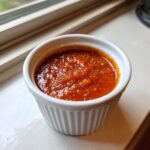 A close-up of a small, white, fluted ramekin filled with bright red, chunky homemade cocktail sauce.