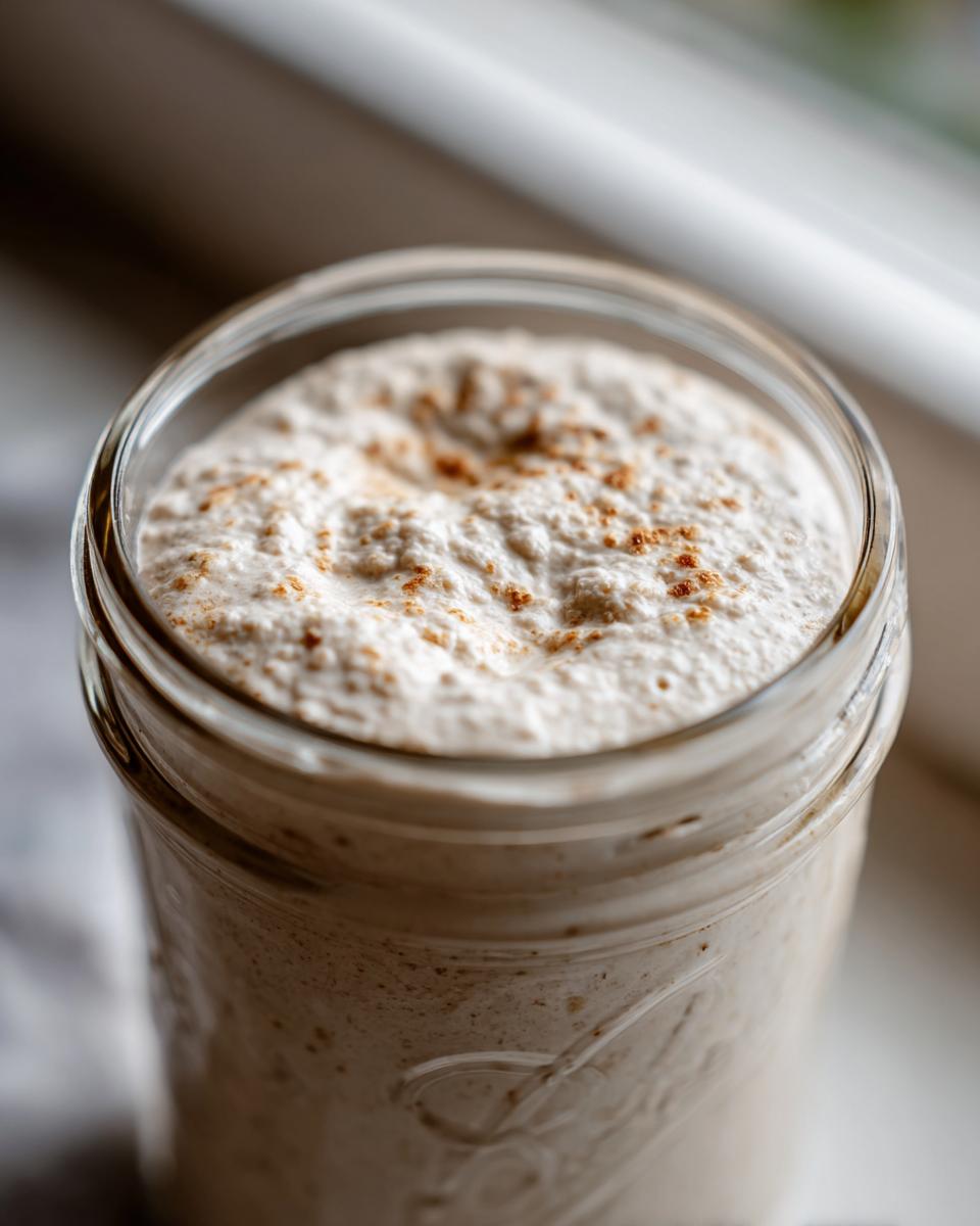 Close-up of a bubbly, active sourdough starter in a glass jar, showing a significant rise and light dusting on top.