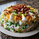 A tall, layered salad featuring visible layers of lettuce, peas, creamy dressing, topped with shredded cheese, hard-boiled eggs, and bacon bits for the perfect 7 layer salad.