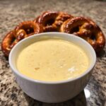A bowl of creamy, yellow beer cheese dip served with soft, salted pretzels in the background.