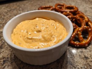 A close-up of creamy, orange beer cheese dip in a white bowl served next to hard pretzels.