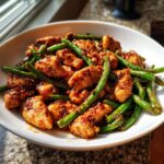 Close-up of savory chicken and green beans coated in a dark, glossy sauce, served in a white bowl.
