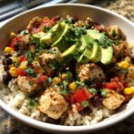 A close-up of a vibrant chicken burrito bowl featuring seasoned chicken, rice, black beans, corn, salsa, and sliced avocado.