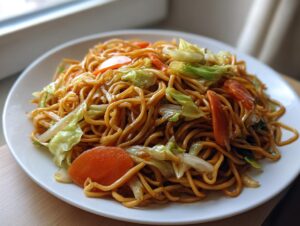 A close-up of a white plate piled high with glossy, savory fried noodles mixed with sliced carrots and cabbage.
