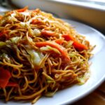 A close-up shot of a plate piled high with savory fried noodles mixed with bright orange carrots and green cabbage.