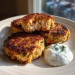 Four perfectly cooked salmon cakes served on a plate with a dollop of creamy dill sauce.