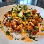 A close-up, appetizing shot of a shrimp bowl featuring grilled shrimp, white rice, corn, avocado, and a creamy orange sauce.