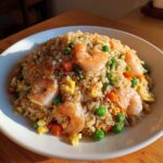 A close-up of a generous serving of shrimp fried rice featuring large pink shrimp, bright green peas, carrots, and scrambled egg.