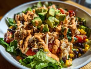 Close-up of a vibrant southwest chicken salad topped with shredded chicken, diced avocado, corn, black beans, and creamy orange dressing.