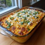 A freshly baked, golden brown street corn dip topped with crumbled white cheese and fresh cilantro in a clear glass baking dish.