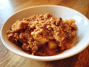 Close-up of a warm serving of apple crumble with a thick, brown, crumbly topping in a white bowl.