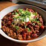 Close-up of a rich bowl of homemade chili topped with sour cream, melted cheese, and fresh green onions.