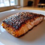 Close-up of a perfectly cooked fillet of blackened salmon with a dark, flavorful crust on a white plate.