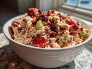 A close-up of creamy blt chicken salad mixed with cherry tomatoes, celery, and topped with crispy bacon bits in a white bowl.