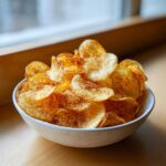 A white bowl overflowing with freshly made, crispy, seasoned potato chips.