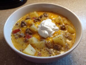 A close-up of a white bowl filled with creamy, cheesy cheeseburger soup featuring chunks of potato, ground beef, and topped with sour cream.