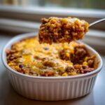 A spoonful of cheesy mexican rice casserole being lifted from a white baking dish.