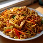 Close-up of a plate piled high with homemade chicken chow mein recipe, featuring noodles, chicken pieces, carrots, and cabbage.