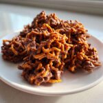 A close-up of several rich, chocolate-coated haystack cookies piled on a white plate.